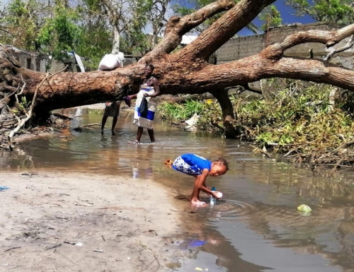 Ein Kind spielt im Wasser nach einer Überschwemmung in Mozambique. Im Hiintergrund ist ein umgefallener Baum. Eine Frau sieht dem Kind zu.