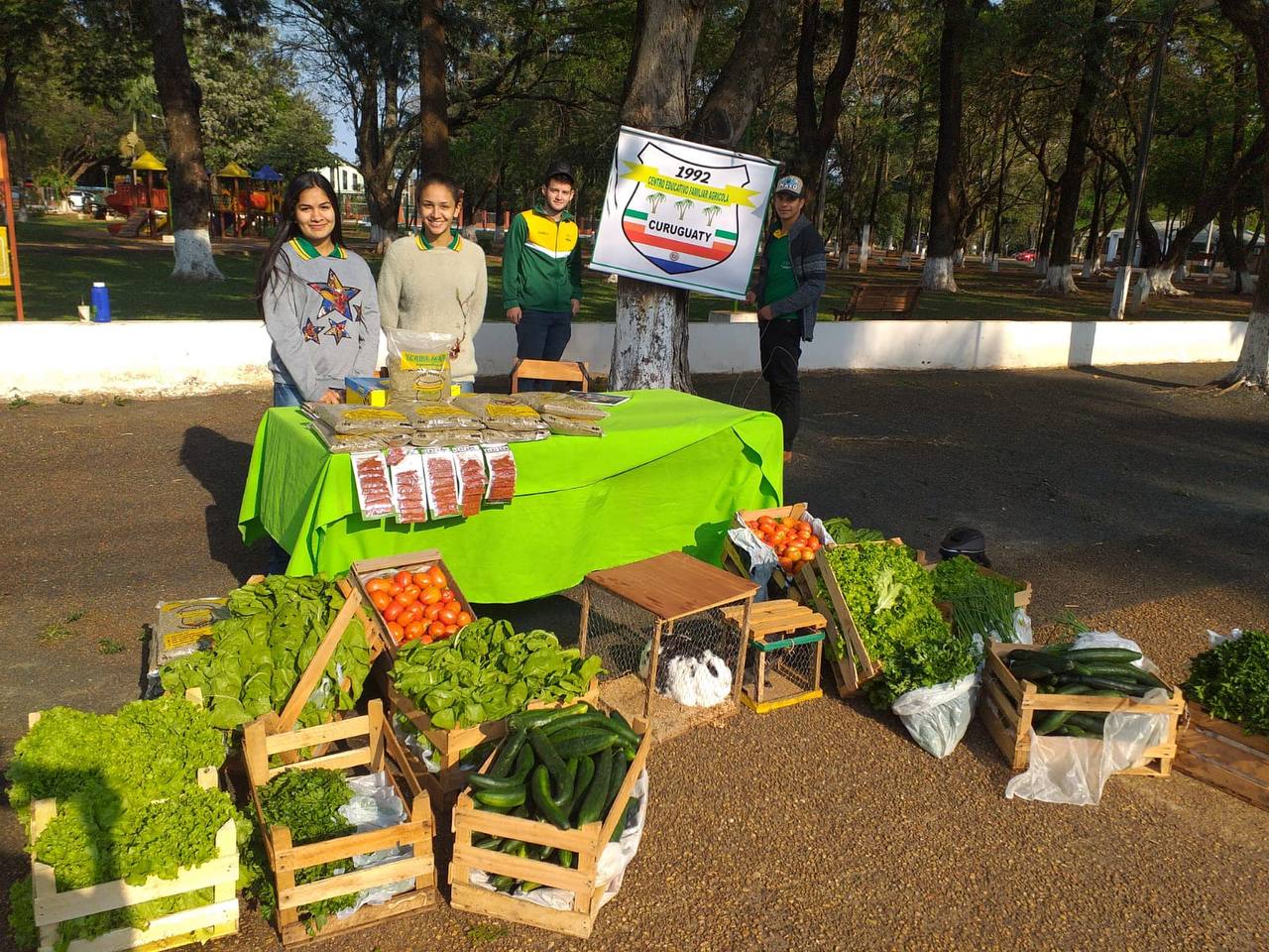 Vier Jugendliche stehen vor einem Stand mit Gemüse, zwei von den Jugendlichen stehen neben der Fane der Schule im Hintergrund