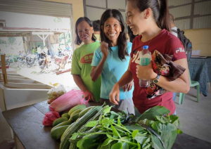 Das Bild zeigt drei lachende Frauen, die vor einem Tisch mit Gemüse stehen.